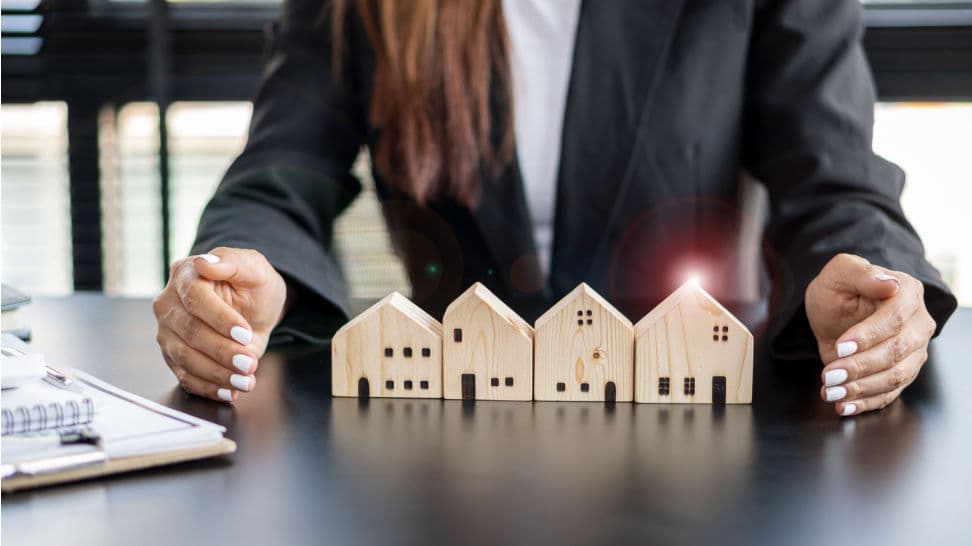 Businesswoman in a dark suit cupping her hands protectively around four small wooden model houses on a desk