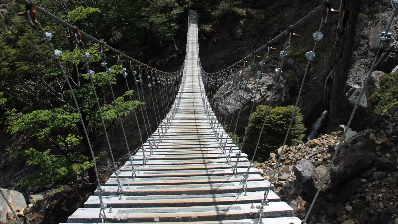 Wooden suspension footbridge stretching across a rocky canyon, with cable supports and weathered slats
