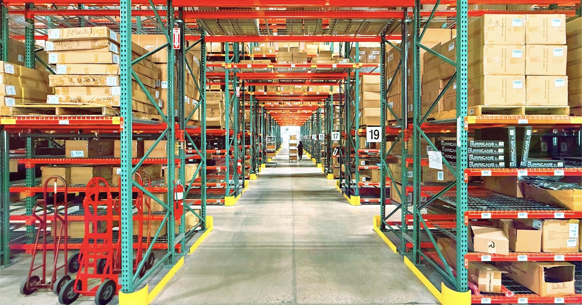 A long aisle between tall steel racks of palletized inventory in a distribution warehouse