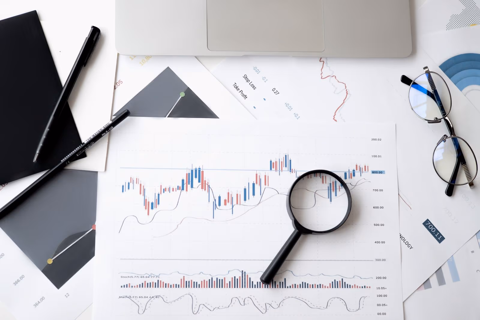 A magnifying glass resting on a printed financial chart surrounded by analysis documents, notes, and reading glasses