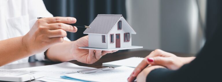 Person holding a model house while another person listens, with documents and a pen on the table